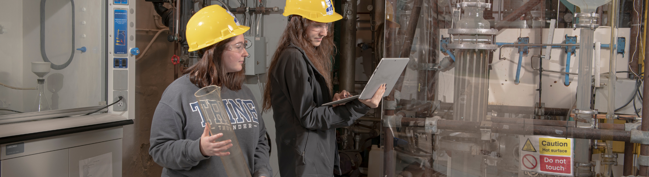 two trine students wearing hard hats and checking equipment in a chemical engineering lab