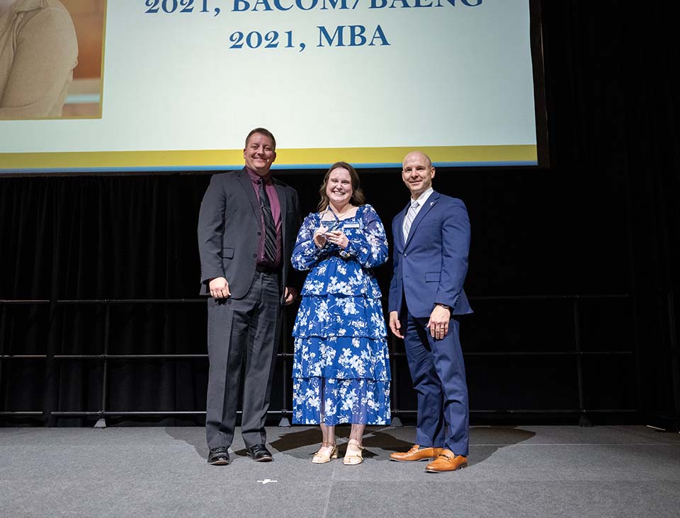 Two men and a woman, all well-dressed, standing on a stage. The woman is in the middle and is holding an award.
