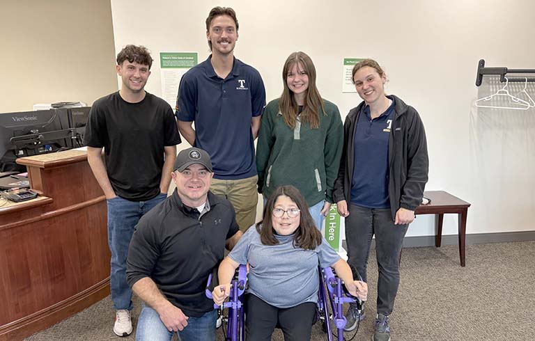 Four Trine students standing behind Sophia Dillon, who is in the pediatric walker they designed, and her father Rob