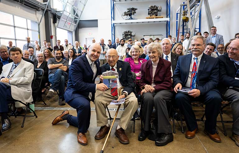 Dr. Kline kneeling as he hands award to John and Mary Pellegrino, with crowd surrounding them.